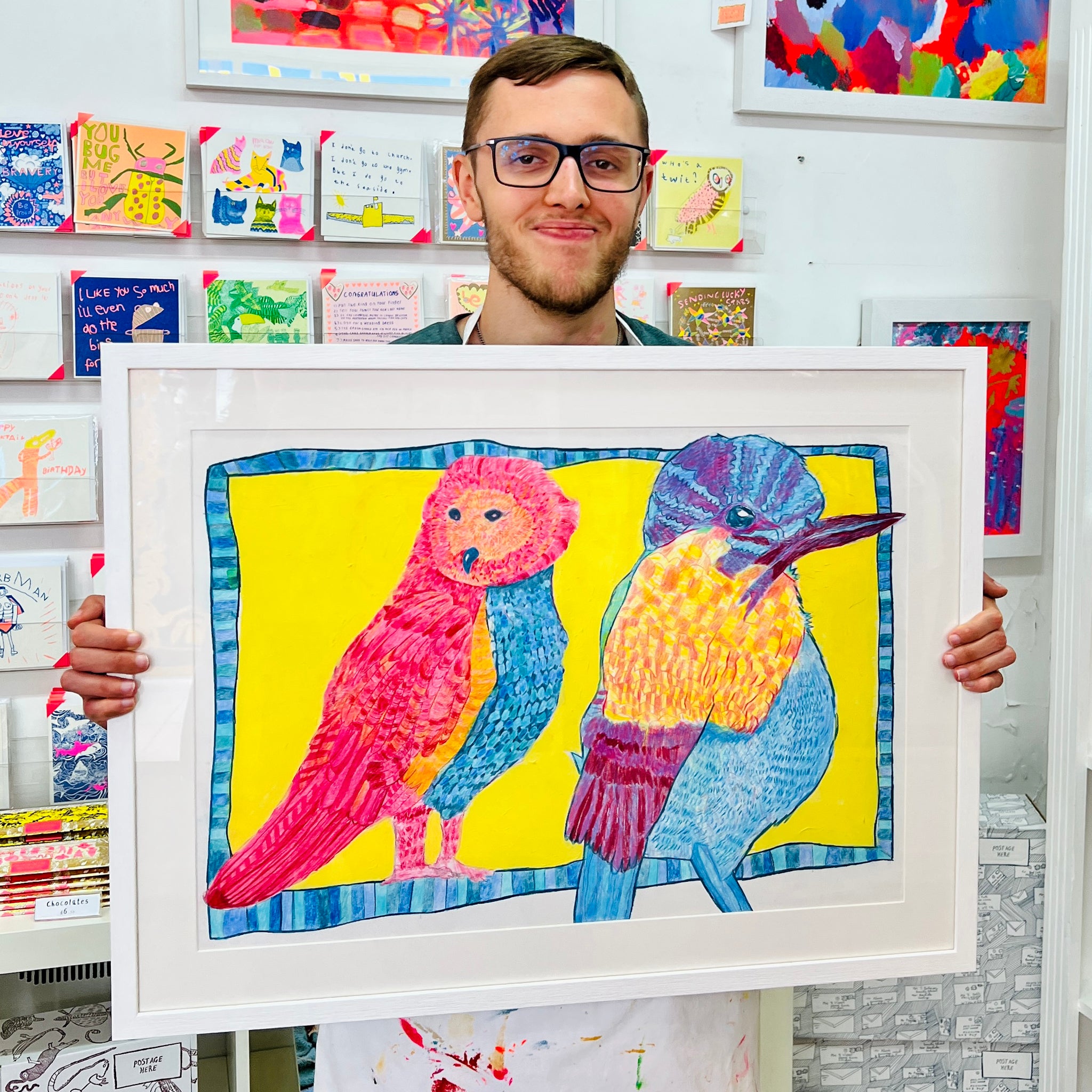 Male artist holding Framed drawing of two pink, yellow and blue birds 