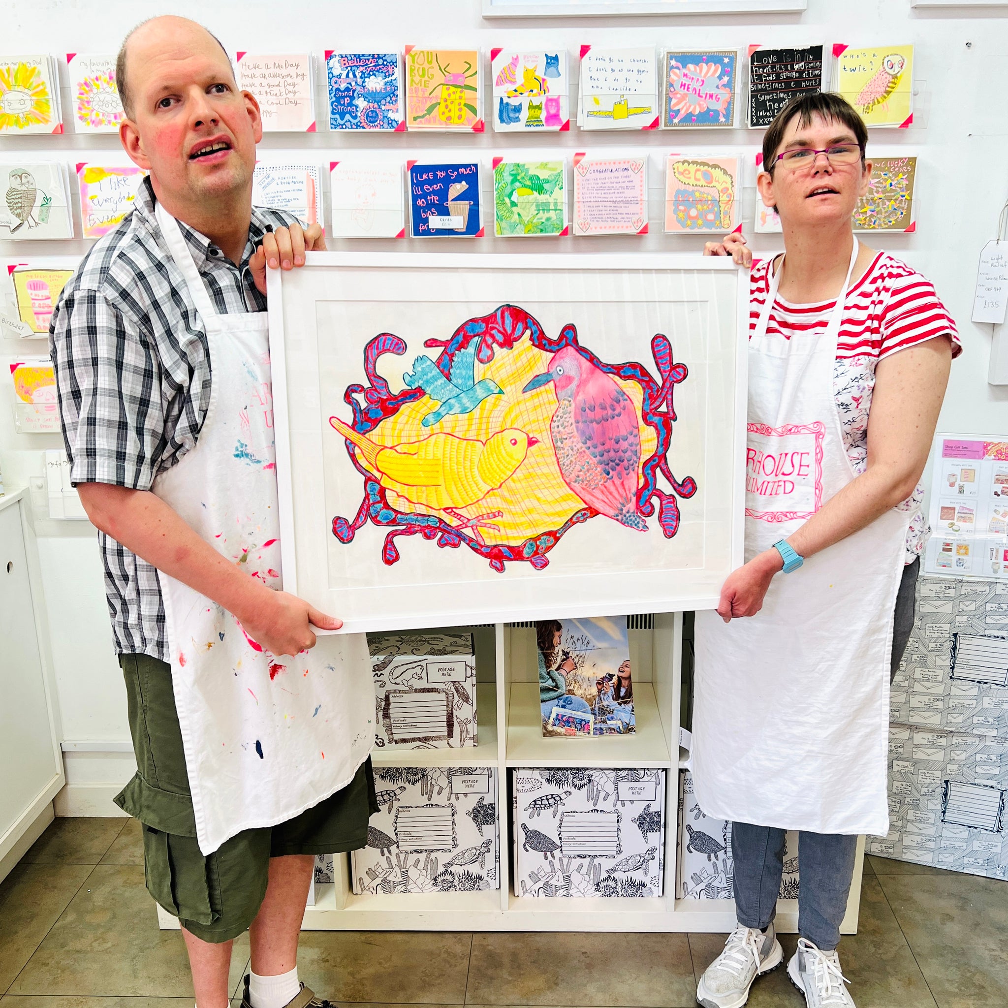 Two artists holding Framed painting of blue, pink and yellow birds 