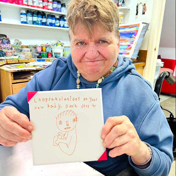 Female artist holding White card with an orange hand drawn image  of a baby with the words Congratulations on your new baby, don't drop it