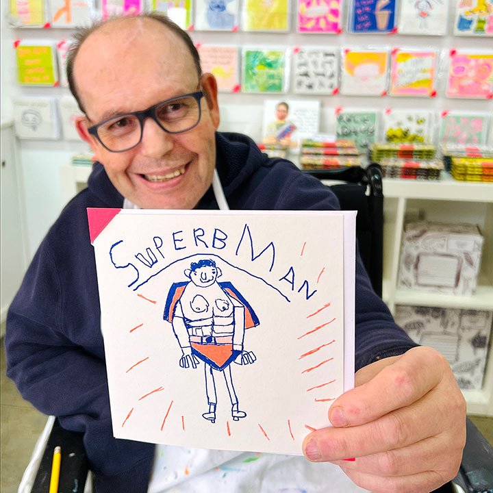 A male artist holding a white greetings card with a hand drawn picture of a man in blue and orange with the words Superb Man 