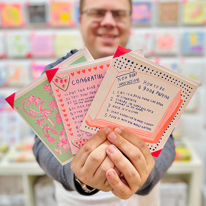 An artist holding 3 cards focusing on A blue and orange card with a hand drawn book with a list of how to be a good parent 