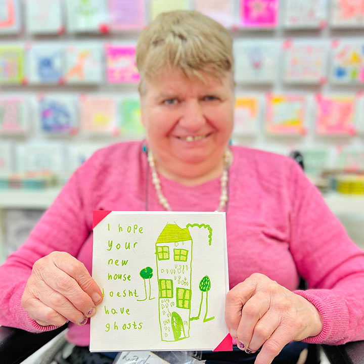 A female artist holding  A white card featuring a green hand drawn image of a house with the words I hope your new house doesn't have ghosts