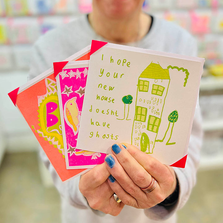 AN artist holding 3 cards focusing on  A white card featuring a green hand drawn image of a house with the words I hope your new house doesn't have ghosts