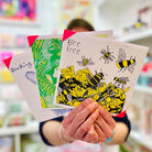 An artist holding 3 cards focusing on White greetings card with a yellow and black hand drawn be design with the words Bee Free