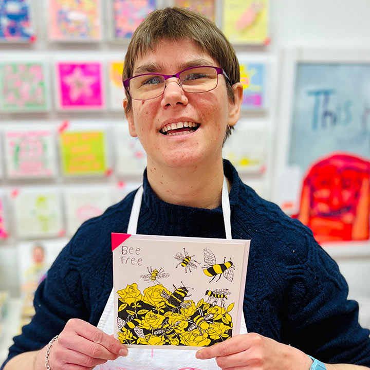 A female artist holding White greetings card with a yellow and black hand drawn be design with the words Bee Free