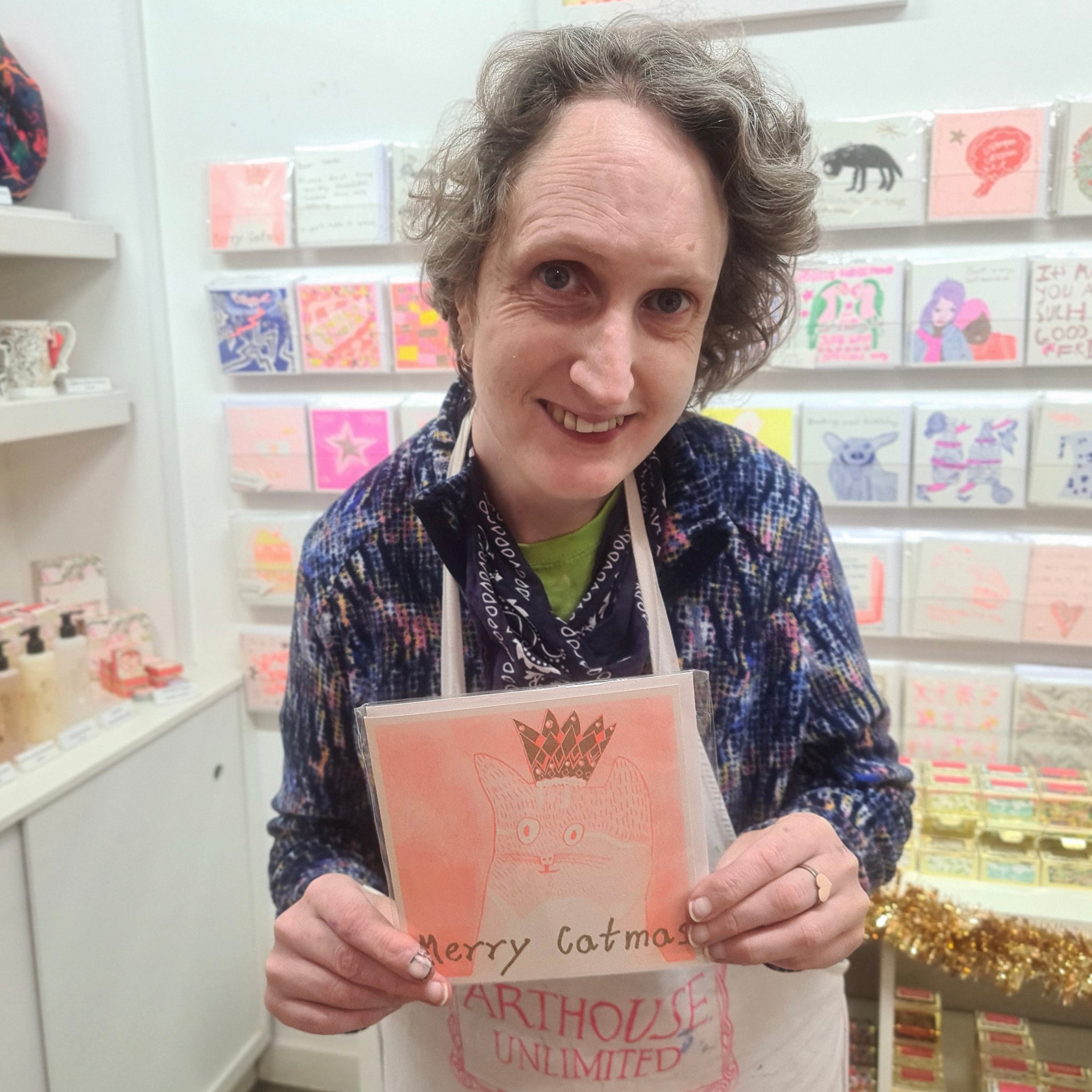 A female artist holding An orange and gold hand drawn christmas card with a picture of a cat and the words merry catmas