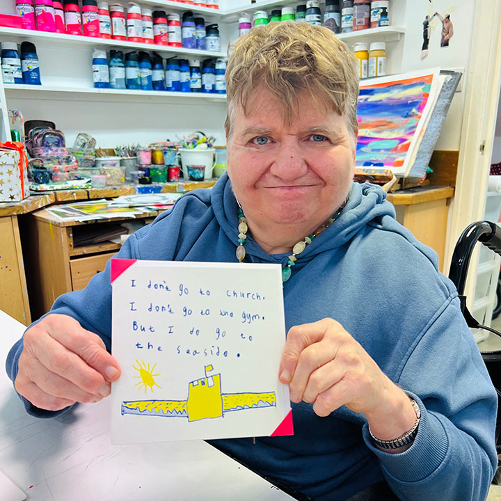 A female artist holding A white card with a blue and yellow drawing of a sand castle with the words I don't go to church, I don't go to the Gym, but I do go to the seaside