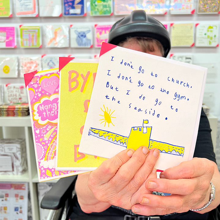 An artist holding 3 cards focusing on A white card with a blue and yellow drawing of a sand castle with the words I don't go to church, I don't go to the Gym, but I do go to the seaside