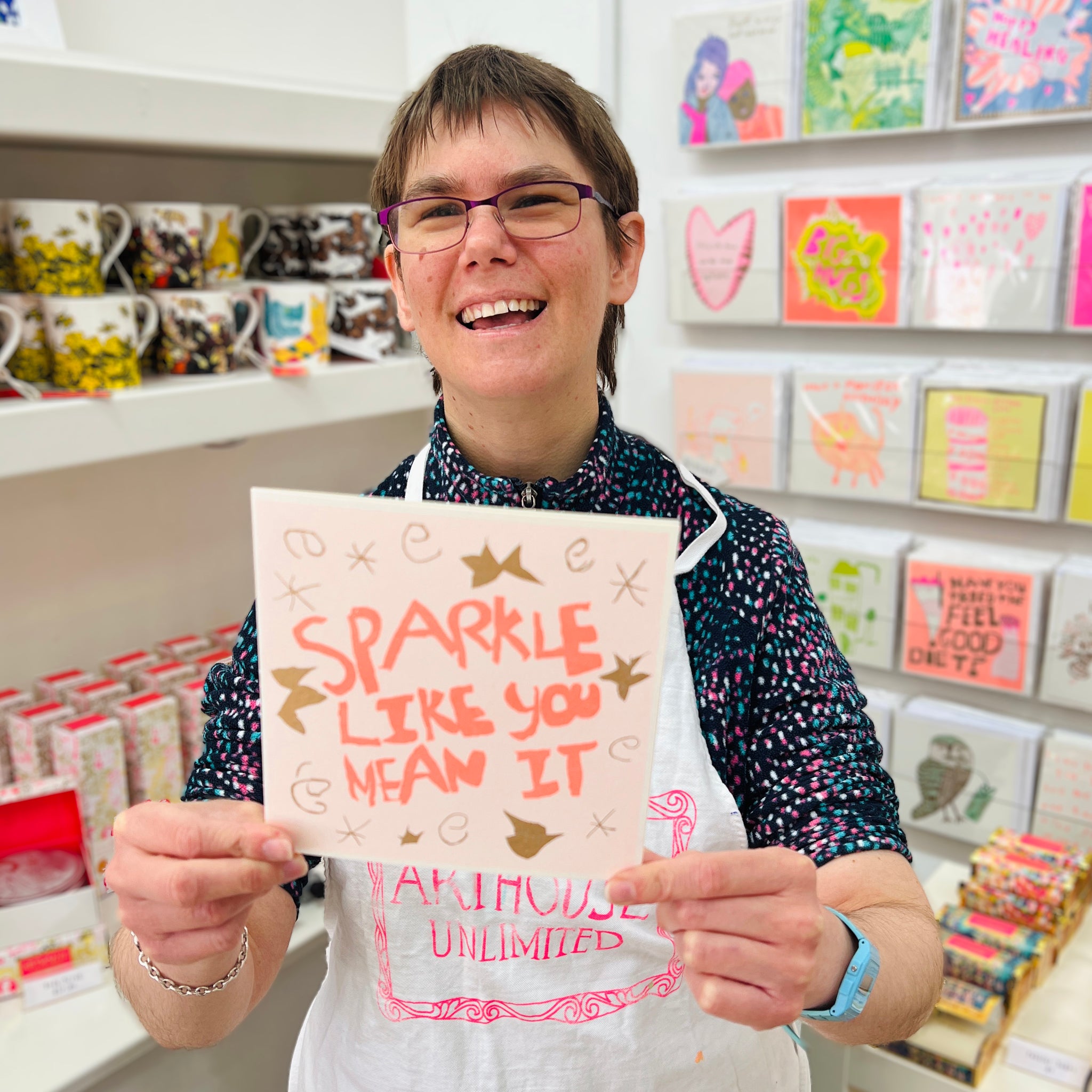 A female artist holding A peach, orange and gold card with the hand drawn words 'sparkle like you mean it' 