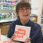 A female artist holding A red, pink and gold hand drawn card with the words merry strange mas