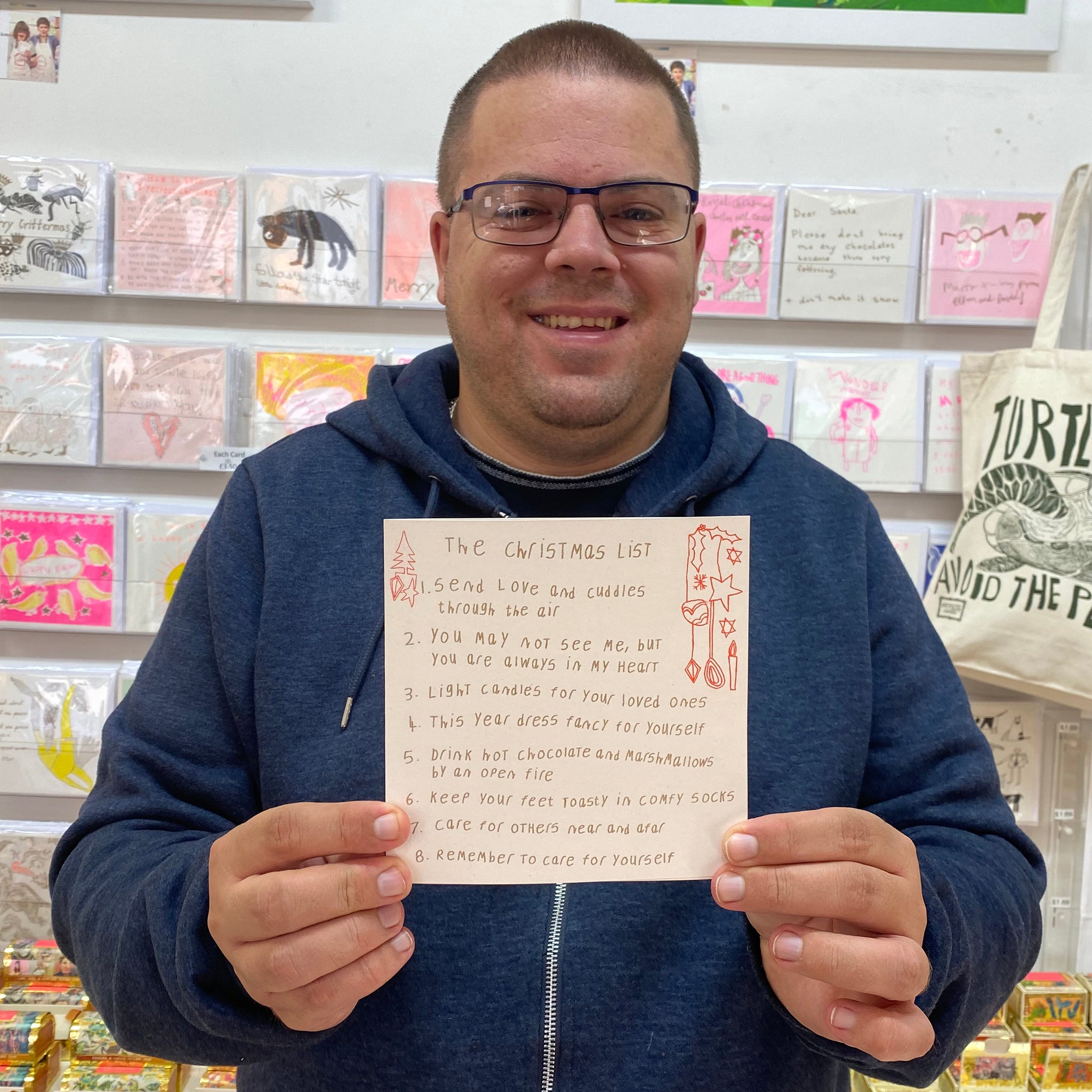 A male artist holding A white card with hand drawn words 'the christmas list' with a list below