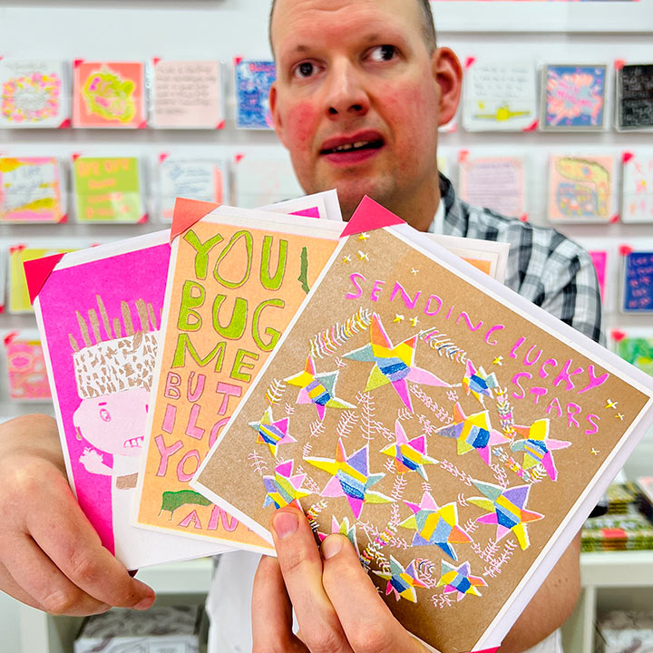 A male artist holding 3 cards focusing on A gold and bright coloured card with hand drawn stars and the words sending lucky stars 
