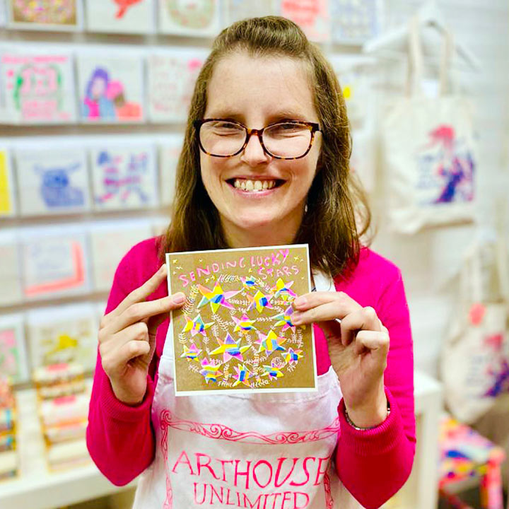 A female artist holding A gold and bright coloured card with hand drawn stars and the words sending lucky stars 