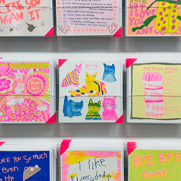 A card rack focusing on A white card with pink, yellow and blue hand drawn cats with the words Miaow for Now