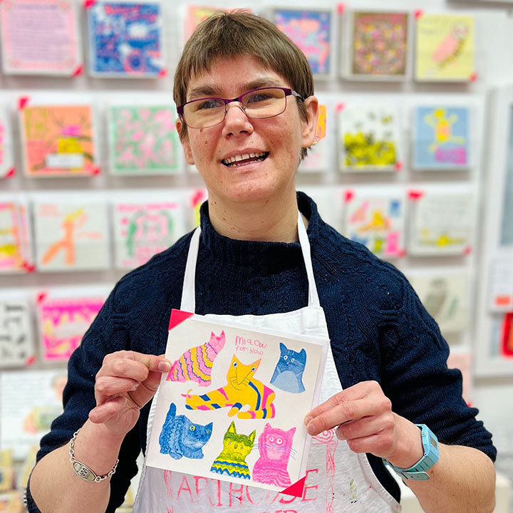 A female artist holding A white card with pink, yellow and blue hand drawn cats with the words Miaow for Now