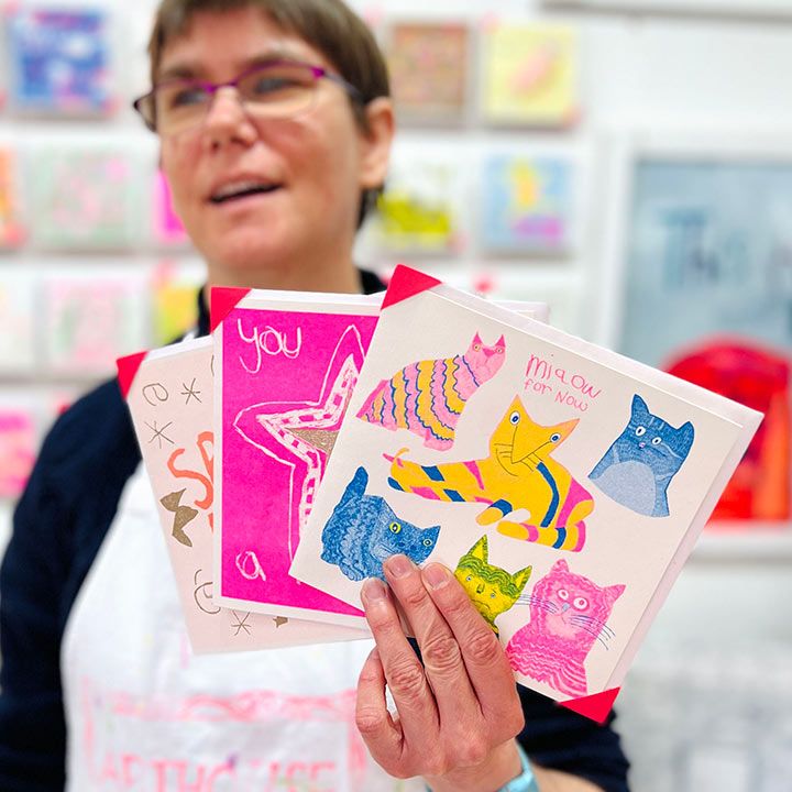 A female artist holding 3 cards focusing on A white card with pink, yellow and blue hand drawn cats with the words Miaow for Now