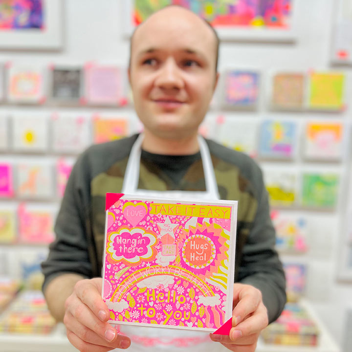 A male artist holding A bright pink, orange and yellow card with positive messages including the words take it easy