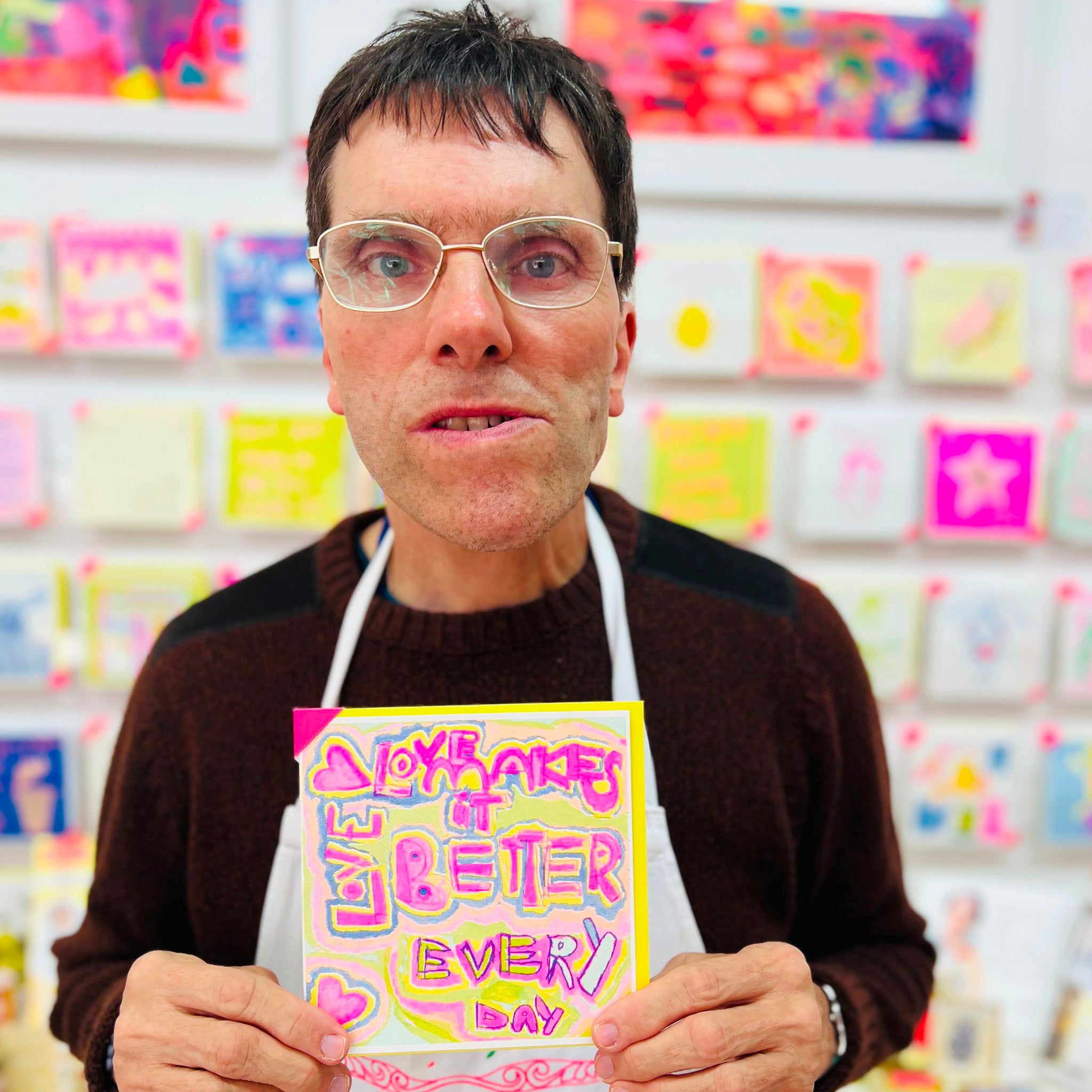 A male artist holding A  bright coloured hand drawn card with the words love makes it better every day 
