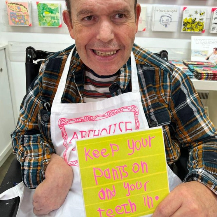 A male artist holding A green card with hand drawn pick words 'keep your pants on and your teeth in