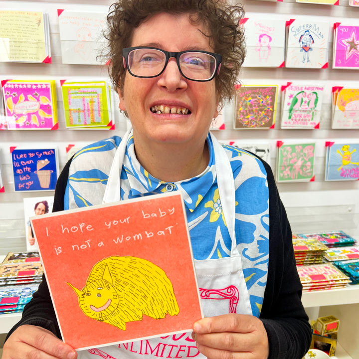 A female artist holding An orange and yellow card with a drawing of a wombat and the words I hope your baby isn't a wombat 