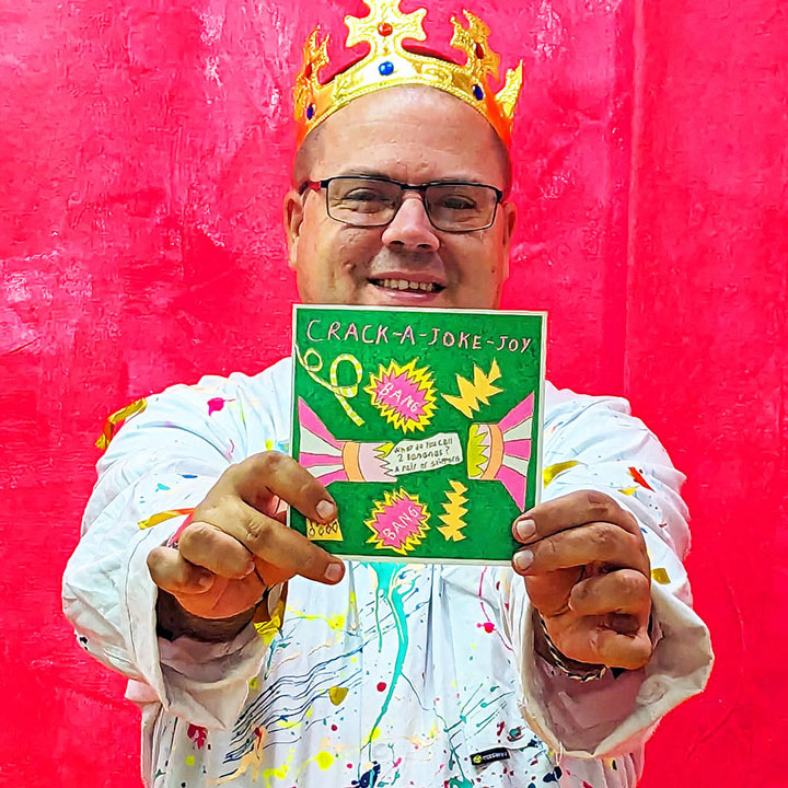 A male artist holding A green, pink and yellow card with a hand drawn cracker and the words crack a joke joy