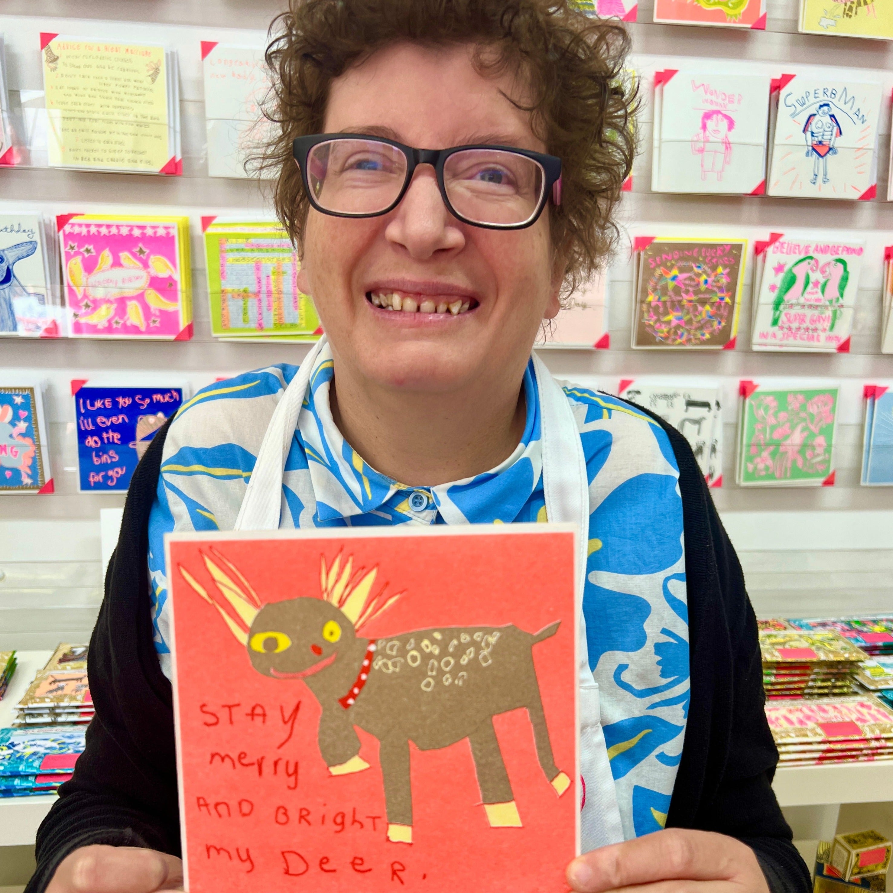 A female artist holding An orange, gold and yellow card with a hand drawn deer and the words stay merry and bright my deer