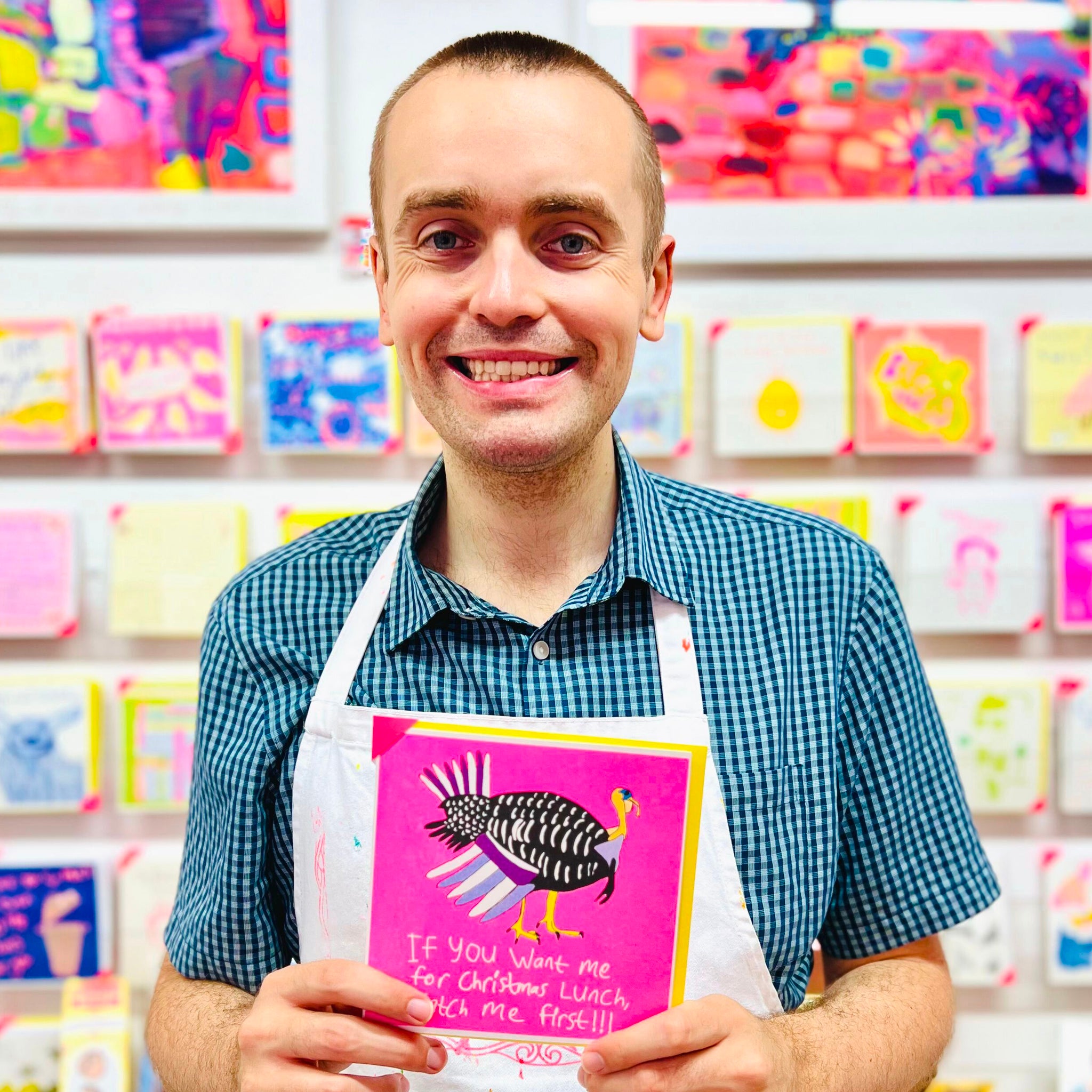 A male artist holding A pink, black and yellow christmas card with a hand drawn picture of a turkey and the words if you want me for christmas lunch, catch me first!!!