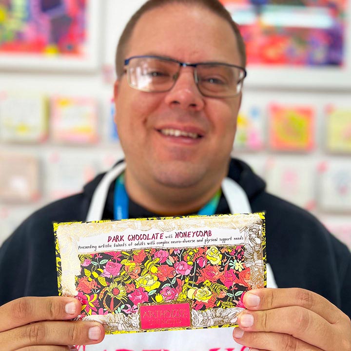 Male artist holding pink, orange, yellow and gold packet of Bee Free, Dark Chocolate Bar with Honeycomb