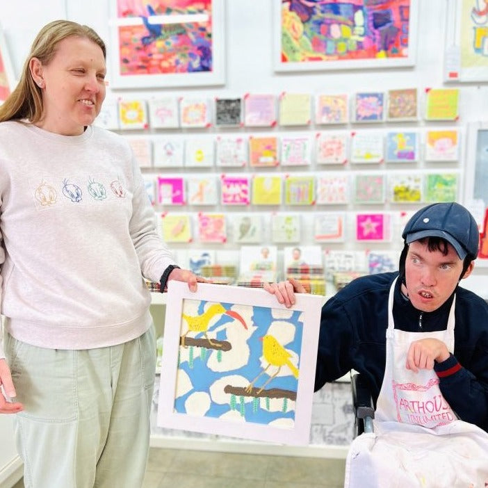 Two artists holdingFramed painted birds in blue and yellow called Morning Gossip