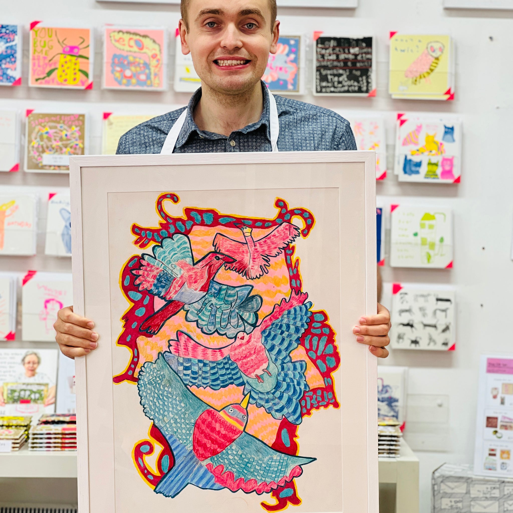 Male artist holding Framed drawing of four blue, pink and yellow birds 