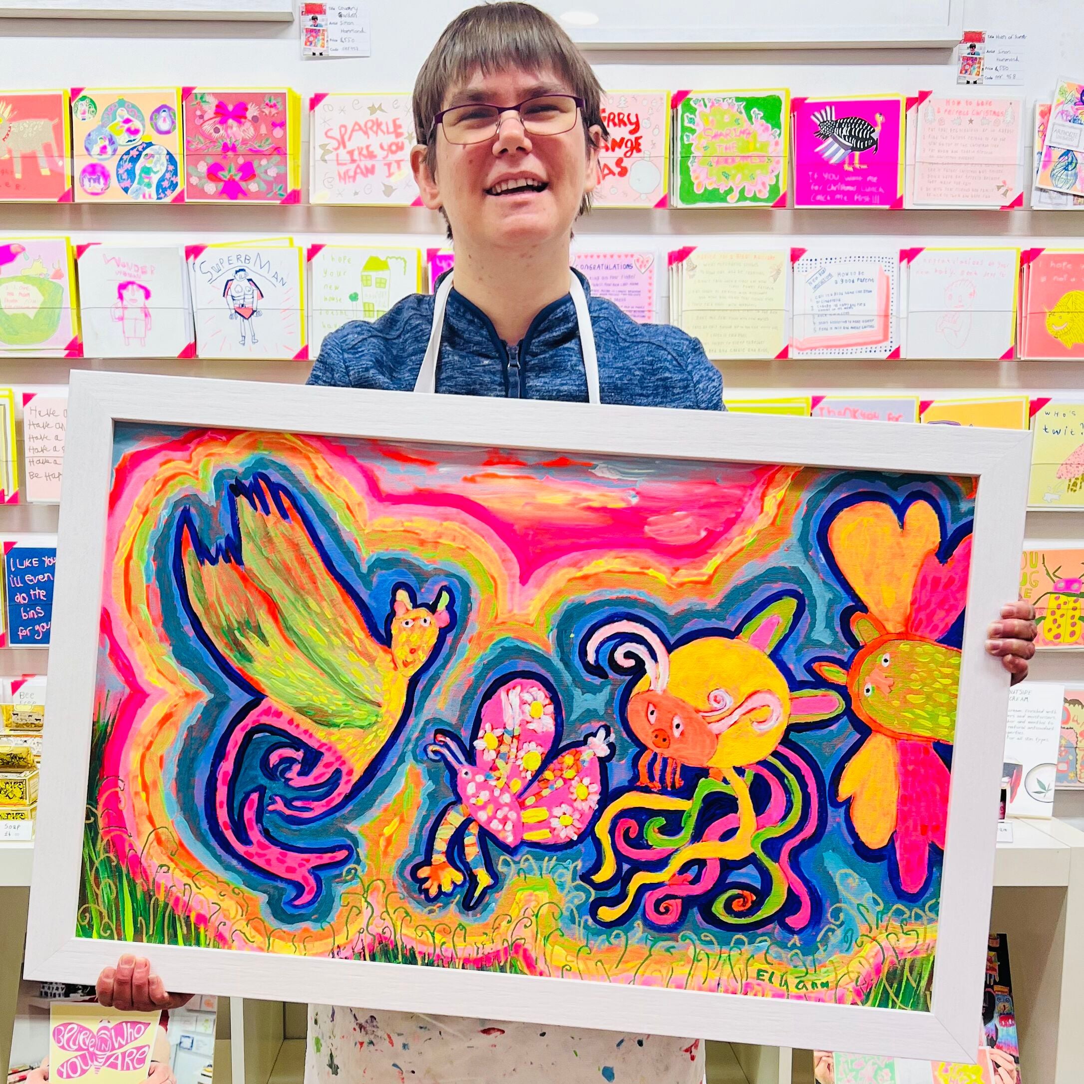 Female artist holding A framed bright coloured painting of strange creatures called Flying Fun with the Friends in the Field