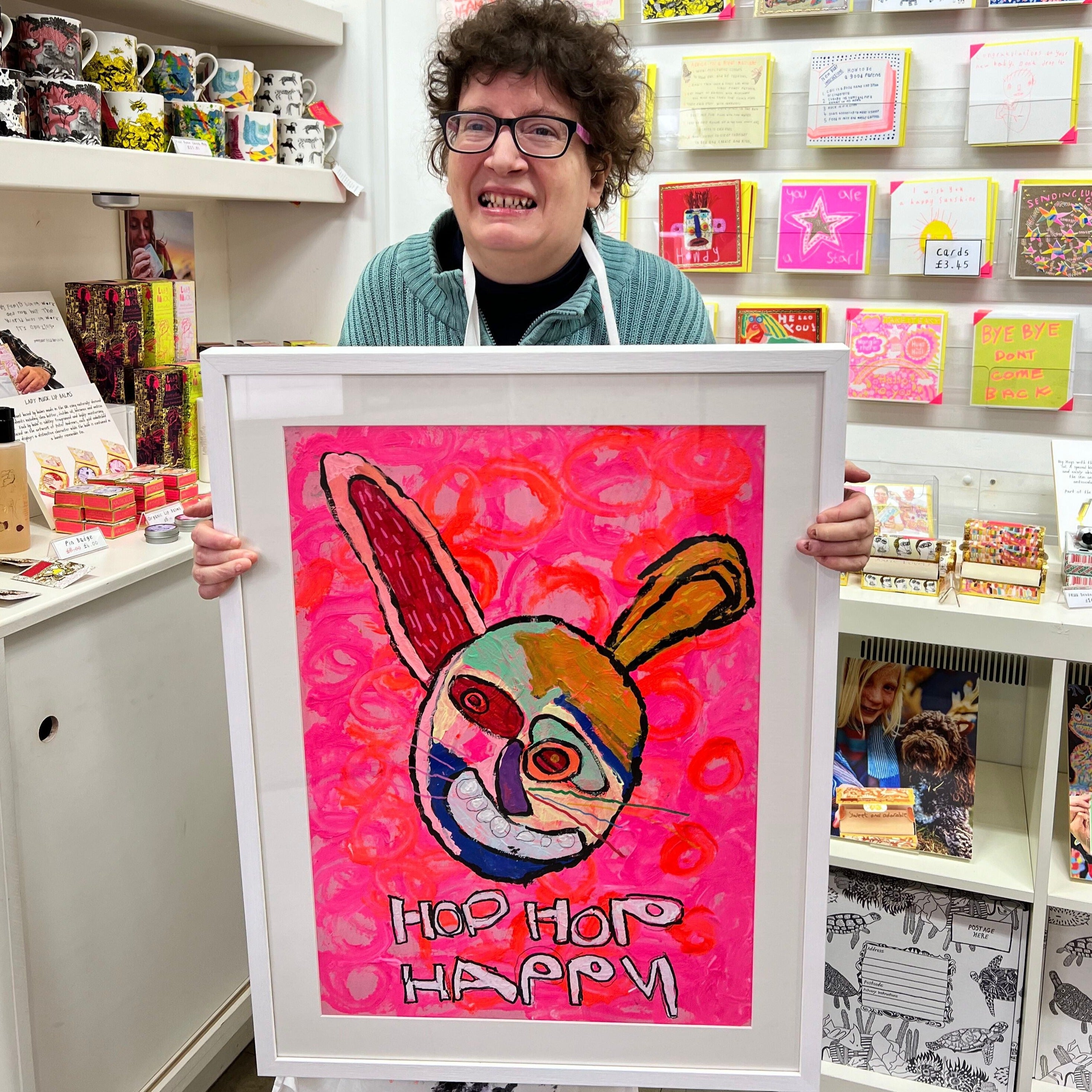 female artist holding Framed painting of a bright coloured rabbit in pinks, red, green and orange with the words hop hop happy