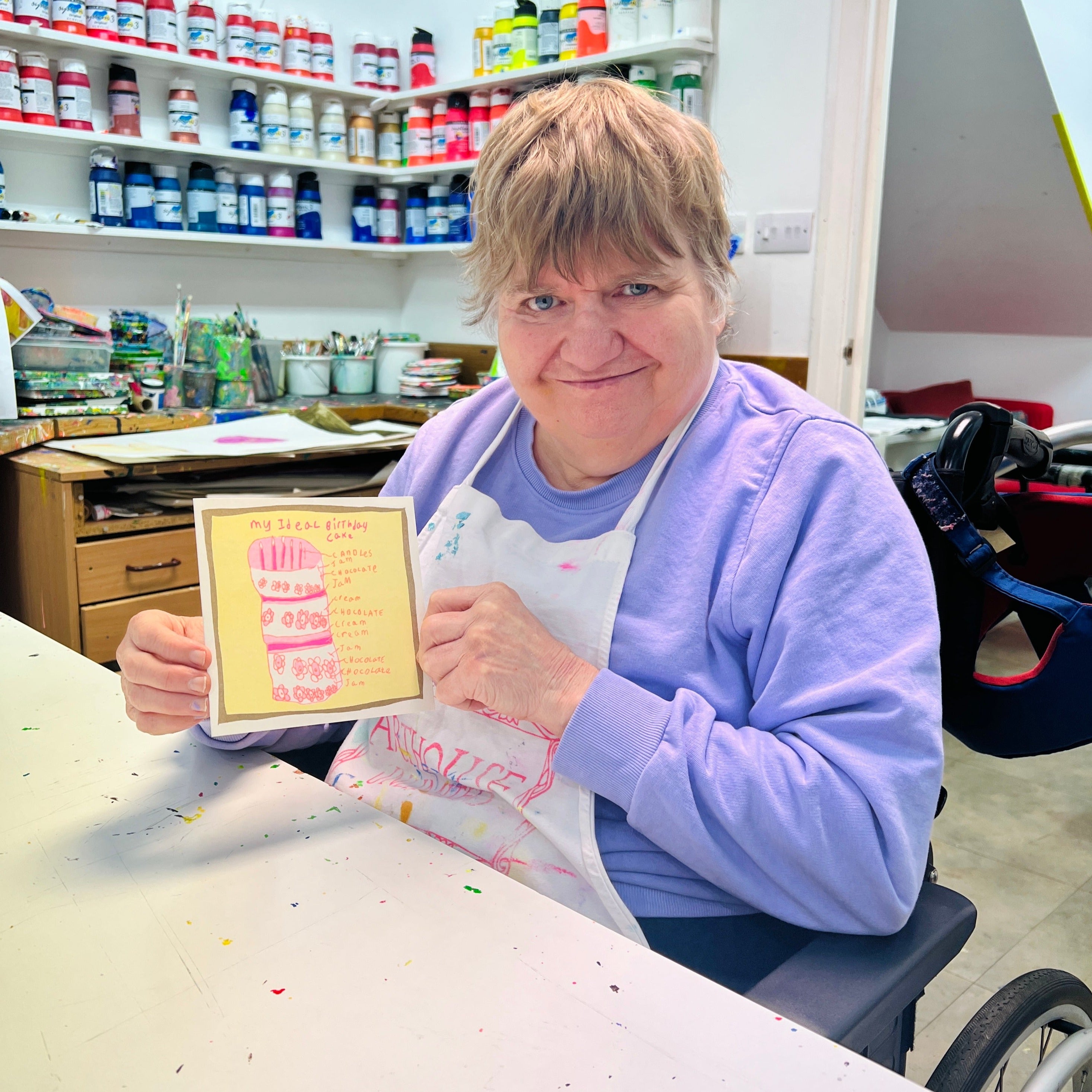A female artist holding A pink, yellow and gold card with a hand drawn birthday cake and the words my ideal birthday cake 
