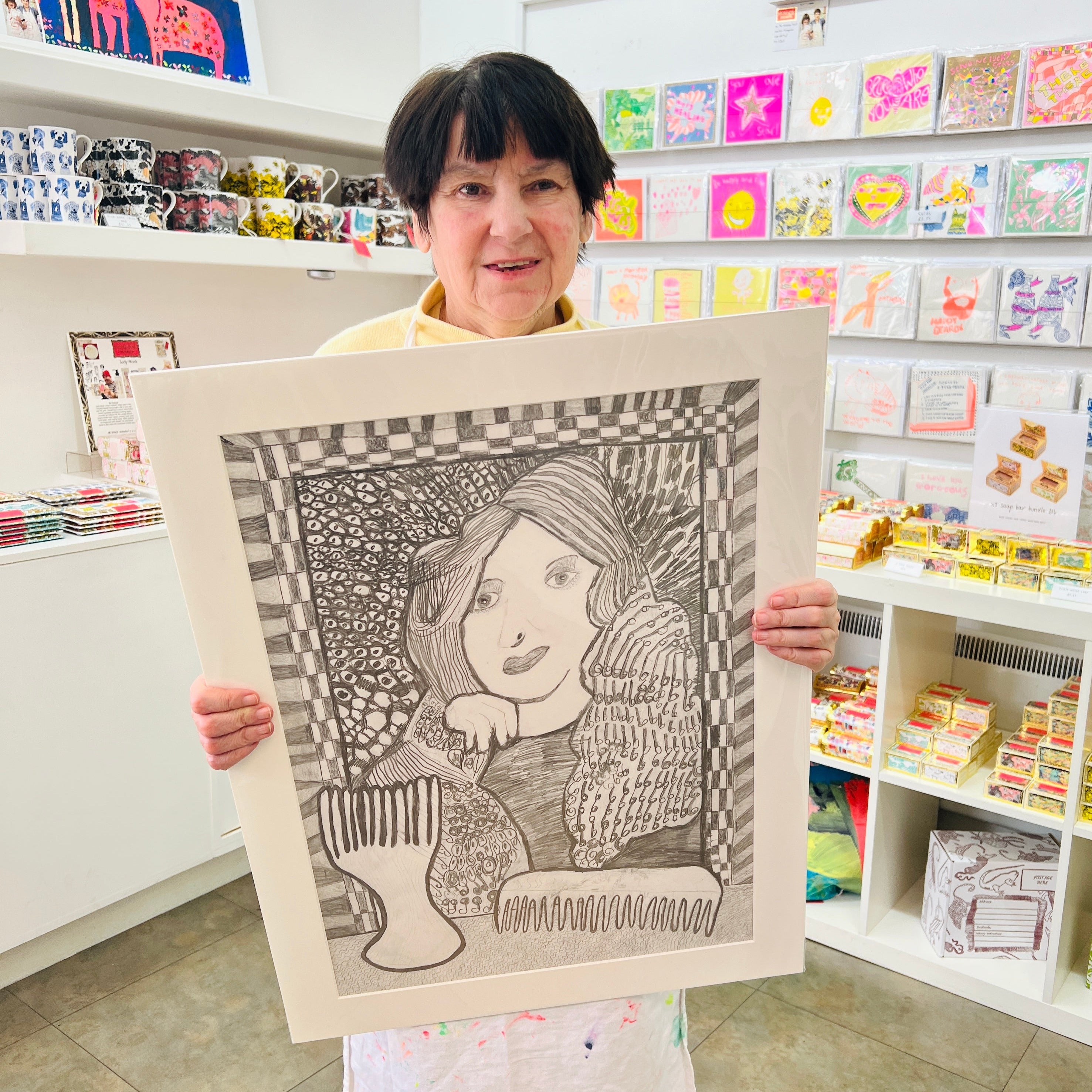 Female artist holding A hand drawn picture in pencil of a woman in front of a mirror with two hair brushes