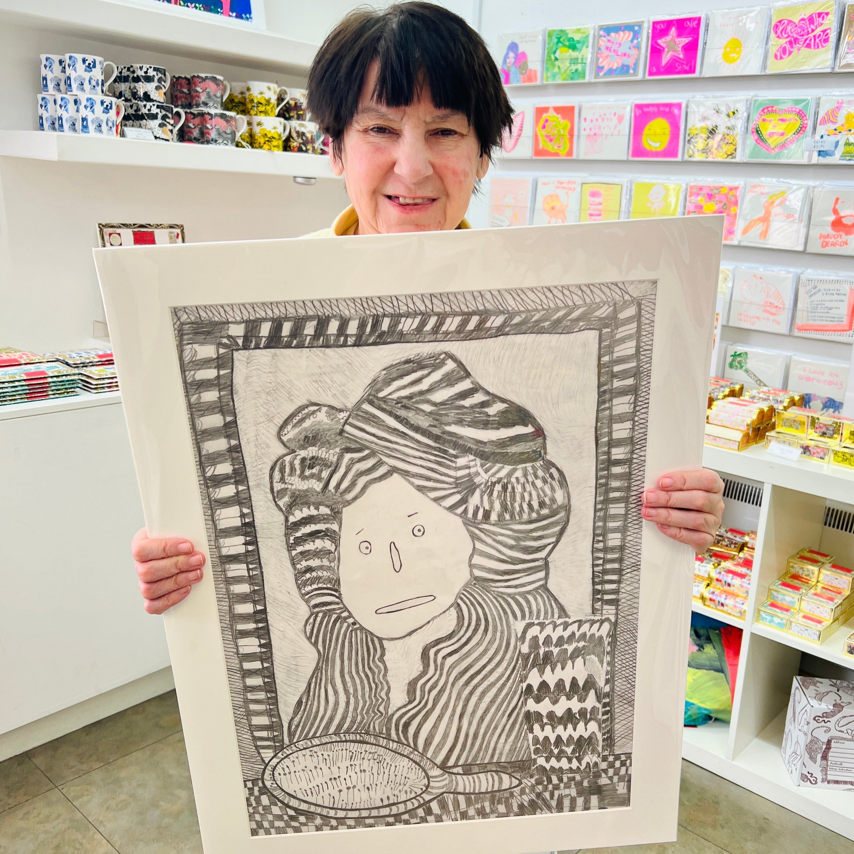 Female artist holding Hand drawn picture in pencil of a woman with a hat on in front of a mirror 