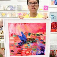 Male artist holding Framed bright coloured hand painted picture of a hummingbird called Hum of Summer