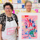 Two artists holding Framed painting of four bright coloured animals stacked on to of one another 