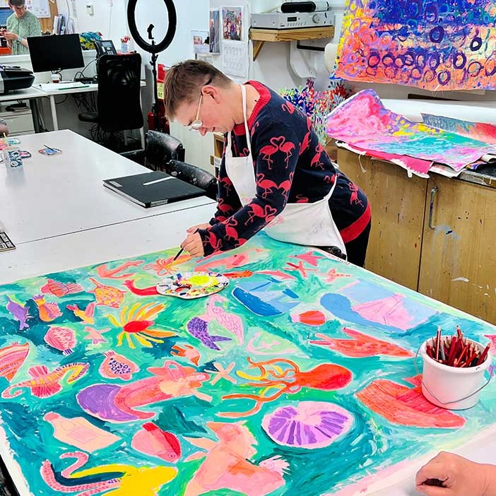 Artist painting on a Large bright coloured painting of mermaids, ships and underwater creatures in blues, oranges and pinks 
