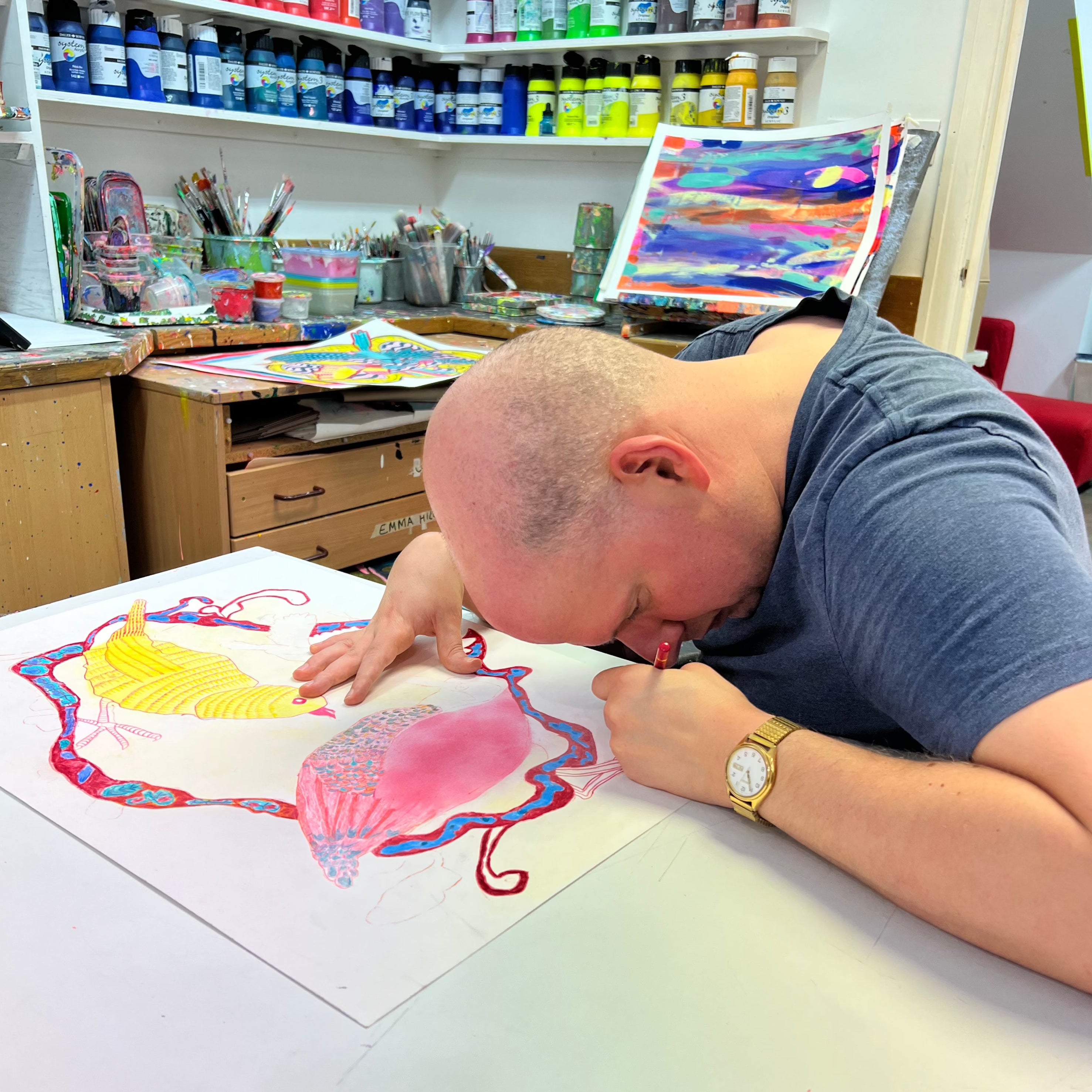 Male artist in a studio drawing bright coloured birds in pencil 