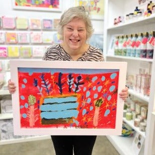 Female artist holding Framed painted artwork of a celebration cake in reds and blues called Celebrate and Decorate