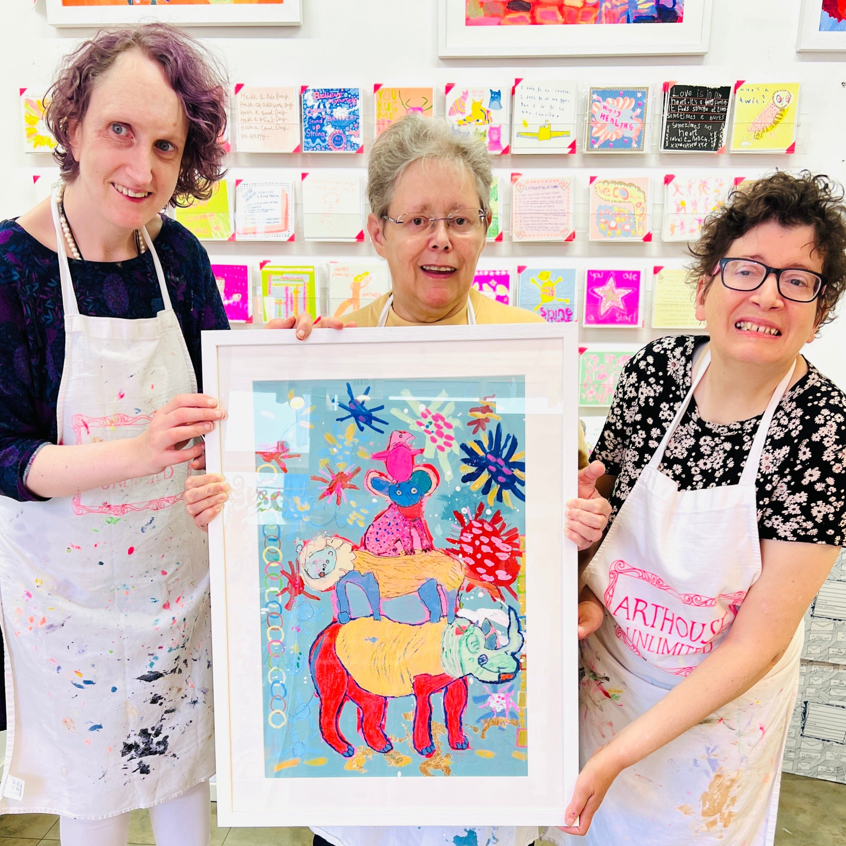Three artists holding Framed painting of four bright coloured animals stacked on to of one another 