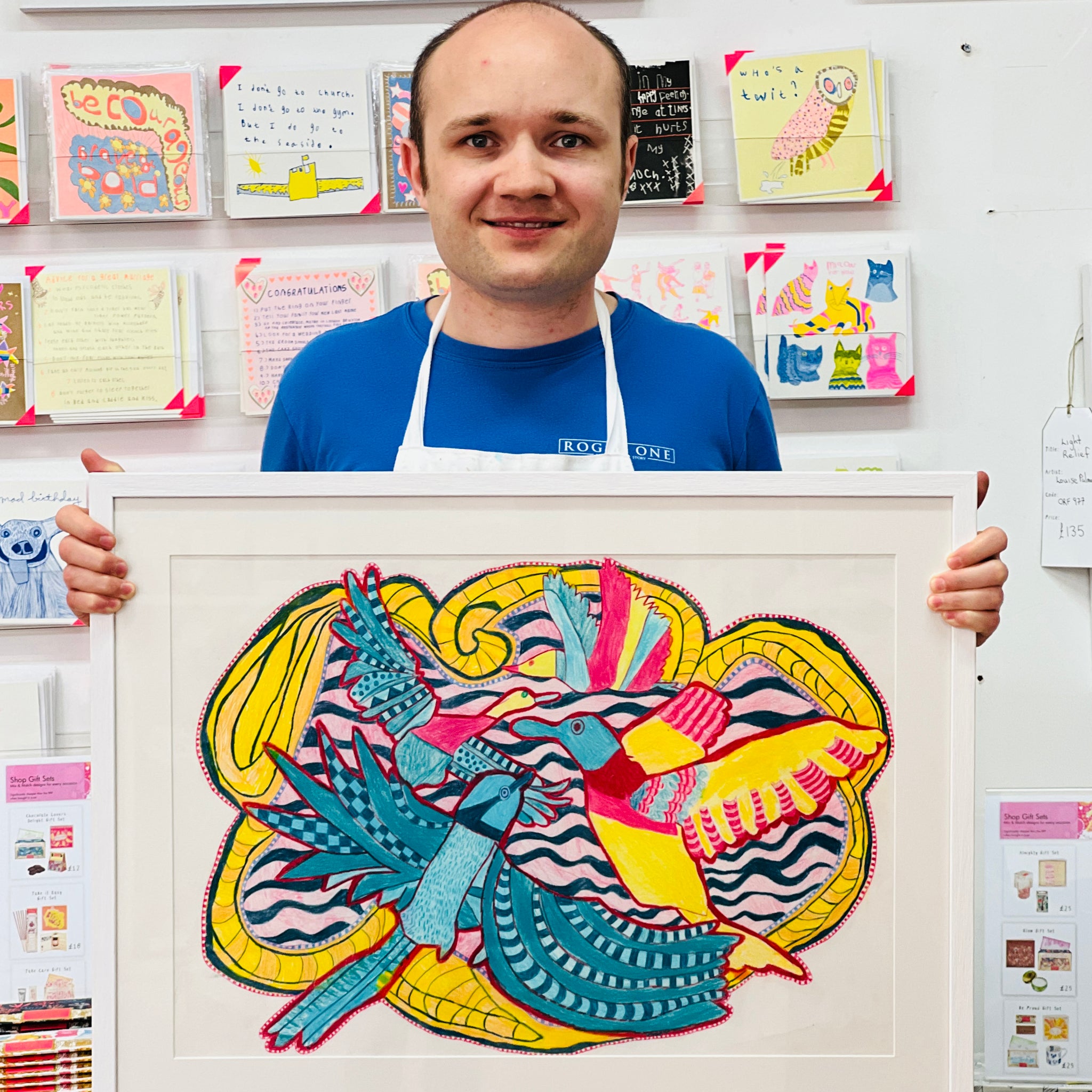 Male artist holding Framed drawing of four blue, yellow and pink birds 