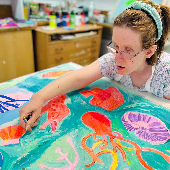 Artist drawing on a Large bright coloured painting of mermaids, ships and underwater creatures in blues, oranges and pinks 