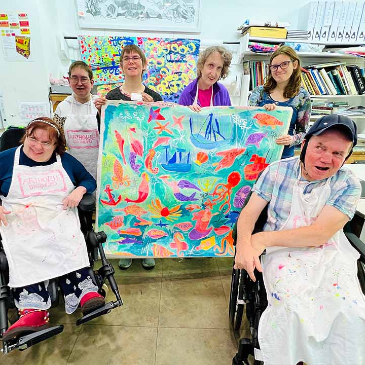 A group of artist holding Large bright coloured painting of mermaids, ships and underwater creatures in blues, oranges and pinks 