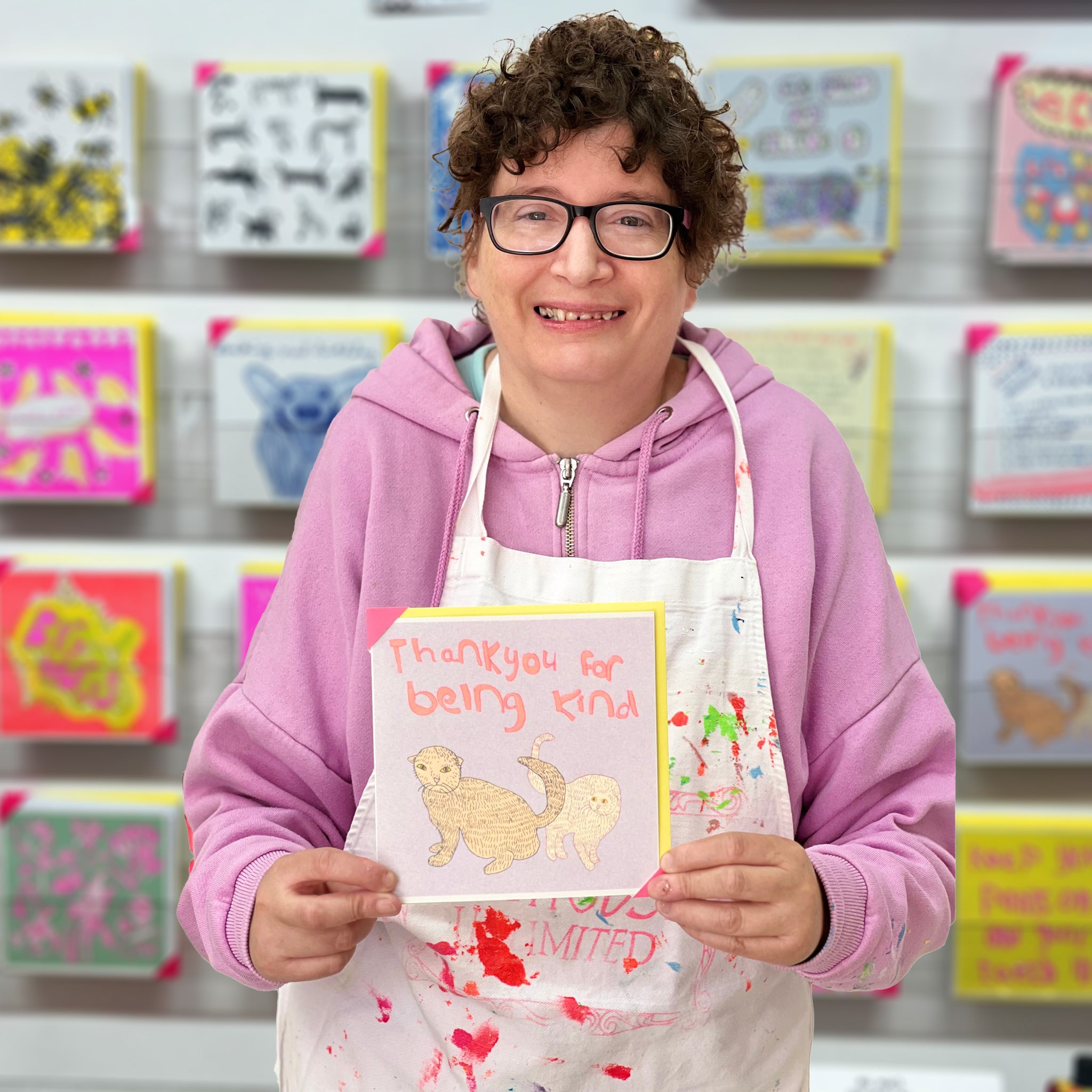 A female artist holding A light purple and orange card with two hand drawn cats and the words thank you for being kind 