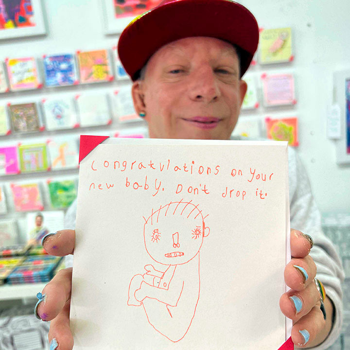 Male artist holding White card with an orange hand drawn image  of a baby with the words Congratulations on your new baby, don't drop it