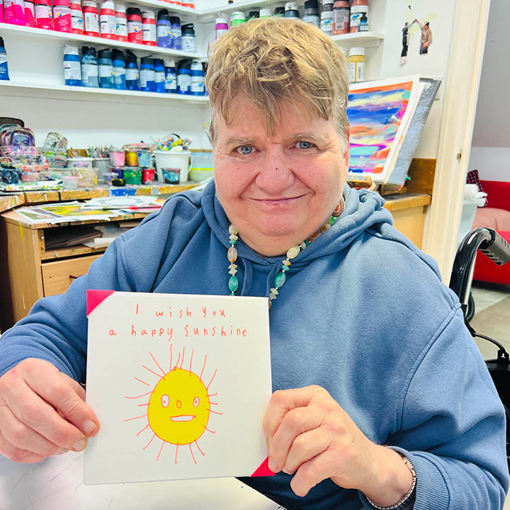 A female artist holding A white card with a hand drawn yellow and orange sun with the words I wish you a happy sunshine