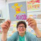 A female artist holding A blue pink and yellow hand drawn character with the words smile inside shine outside