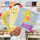 An artist holding 3 cards focusing on A blue pink and yellow hand drawn character with the words smile inside shine outside