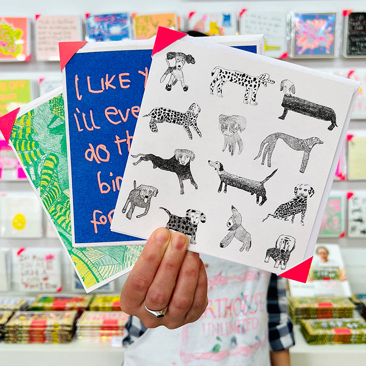 An artist holding 3 cards focusing on A white card with 13 black hand drawn dogs 